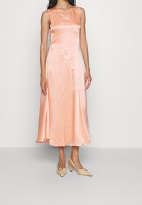 Satin, ärmlös, ankellång klänning i ljus persika med en subtil framklippning och draperad design. Kombinerad med beigefärgade slingback-klackar.