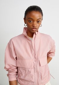 Lys rosa zip-up jakke med høy krage, to frontlommer og svakt rynkete ermer. Glatt, teksturert stoff med en avslappet passform.