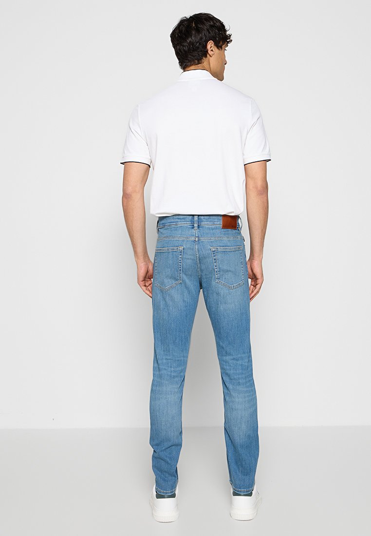 Jean en denim bleu clair avec une coupe slim, comportant des poches arrière et une étiquette en cuir marron à la taille, associé à un polo blanc.