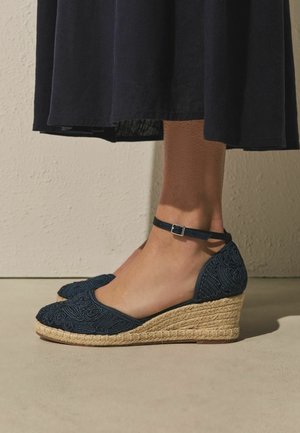 Füße, die marineblaue, bestickte Keil-Espadrilles mit Knöchelriemen tragen, kombiniert mit einem schwarzen Midirock vor einer beigen Wand und einem beigen Boden.