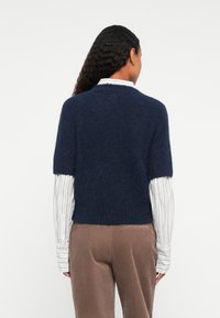 Egy navy kék, rövid ujjú, bolyhos pulóvert viselő személy, alatta egy fehér csíkos, hosszú ujjú inggel és barna kordbársony nadrággal, hátulról látható.