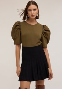Top verde oliva con mangas abullonadas, falda negra con pliegues, textura de tela suave y detalles brillantes en el hombro.