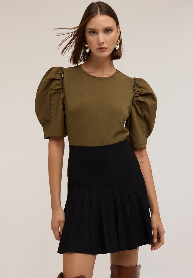 Top verde oliva con mangas abullonadas, falda negra con pliegues, textura de tela suave y detalles brillantes en el hombro.
