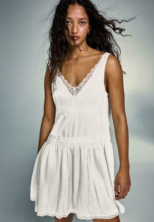 Femeie cu păr creț purtând o rochie albă fără mâneci cu margine din dantelă, stând în fața unui fundal gri neutru.