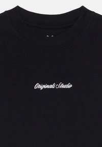 Melnas kokvilnas T-krekls ar apaļu kakla izgriezumu, kuram krūtī ir balti izšūti burti "Originals Studio". Mīksta tekstūra.
