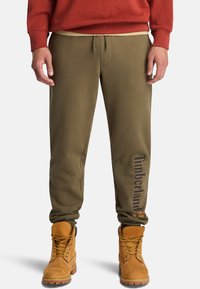 Eine Person steht in olivgrünen Jogginghosen mit "Timberland"-Aufdruck am Bein und tanfarbenen Arbeitsschuhen, kombiniert mit einem roten Sweatshirt.