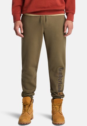 Osoba stoi w oliwkowych dresowych spodniach z napisem "Timberland" na nodze i beżowych butach roboczych, w połączeniu z czerwoną bluzą.