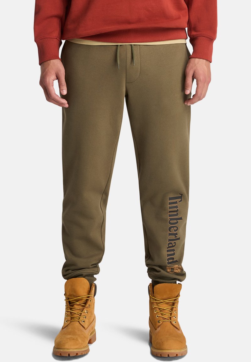 Eine Person steht in olivgrünen Jogginghosen mit "Timberland"-Aufdruck am Bein und tanfarbenen Arbeitsschuhen, kombiniert mit einem roten Sweatshirt.