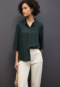 Mörkgrön skjorta med krage och trekvartsärmar, kombinerad med beige byxor. Slätt tyg, enkel design och inga synliga mönster.
