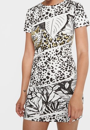 Kobieta w mini sukience z krótkim rękawem w czarno-białe wzory pantery, z dodatkowymi motywami roślinnymi oraz subtelnym wzorem twarzy lamparta na piersi.