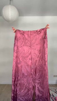 Χέρια κρατούν ροζ σατέν παντελόνια με φαρδιά πόδια τεντωμένα σε έναν απλό τοίχο σε ένα φωτεινό δωμάτιο.