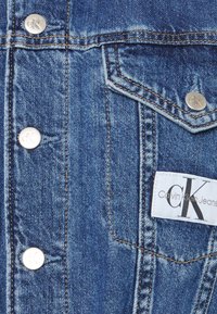 Niebieska dżinsowa kurtka z metalowymi guzikami, przeszyciami oraz białą metką z napisem "Calvin Klein Jeans" i logo.
