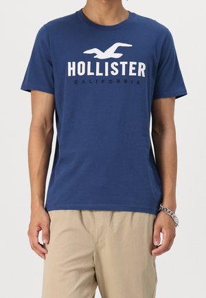 Homme portant un t-shirt bleu Hollister California et un pantalon beige, debout les bras détendus le long du corps.