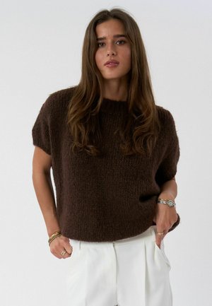 Giovane donna con lunghi capelli castani che indossa un maglione marrone scuro a maniche corte, pantaloni bianchi e braccialetti d'oro e argento, in piedi con le mani nelle tasche.