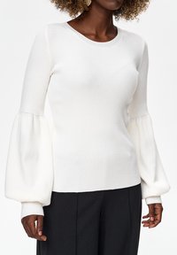 Vrouw draagt een witte geribbelde trui met brede, geplooide ballonmouwen, gecombineerd met een zwarte broek tegen een effen achtergrond.