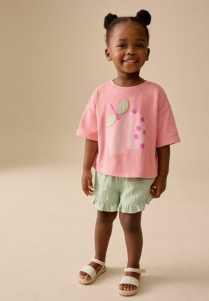 Smiling toddler girl with two puff ponytails wearing a pink shirt with fruit design, light green ruffled shorts, and sandals.
