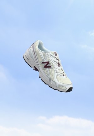 Baskets blanches athlétiques avec un logo "N" bordeaux flottant sur un fond de ciel bleu clair.