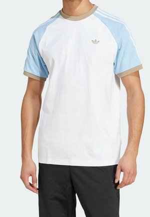 Mann trägt weißes T-Shirt mit hellblauen Ärmeln, beigem Kragen und Bündchen sowie schwarze Hose, steht vor einem schlichten hellen Hintergrund.