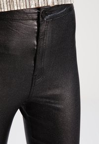 Svarta byxor med metallisk finish och slim fit, med en traditionell knapp- och dragkedjestängning, samt en subtil glans. Detaljsydd synlig.