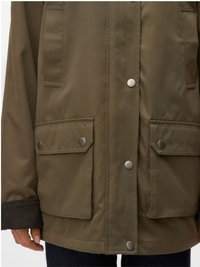 Zielono-oliwkowa kurtka z zatrzaskami, dwoma dużymi kieszeniami z klapami z metalowymi zatrzaskami oraz ciemnymi ściągaczami na rękawach, noszona na ciemnych spodniach.