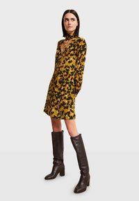 Keltainen mekko, jossa on musta ja ruskea kukkakuosi, pitkähihaiset, syvä V-pääntie, yhdistettynä korkeisiin ruskeisiin nahkabootseihin ja choker-kaulaketjuun.