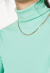Mintgrön ribbad turtleneck med en mjuk textur, prydd med ett tunt guldhalsband med avlånga länkar som bärs över den.