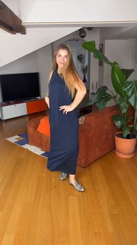 Γυναίκα με μακριά ξανθά μαλλιά, φορώντας αμάνικο μπλε φόρεμα και παπούτσια με σχέδια, ποζάρει σε ένα σαλόνι με μεγάλο φυτό και πορτοκαλί καναπέ.