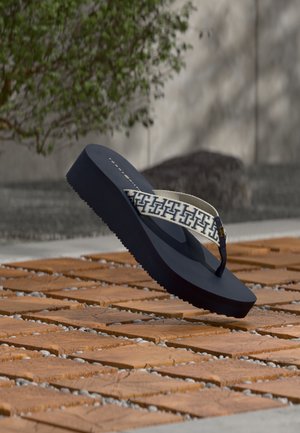 Marineblå platforme flip-flop med en hvid mønstret rem hviler på en udendørs træflise og småsten.