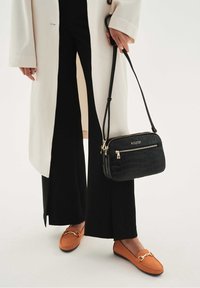 Borsa a tracolla nera in ecopelle con motivo coccodrillo, con hardware dorato, due scomparti con zip, indossata con un cappotto crema e pantaloni neri a gamba larga.