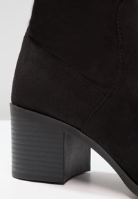 Bottine en suede noir avec un petit talon carré et des détails de couture visibles sur une surface blanche.