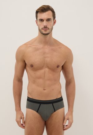 Gespiierde man staande tegen een egale achtergrond, gekleed in grijze slips met een zwarte tailleband en zwarte details.