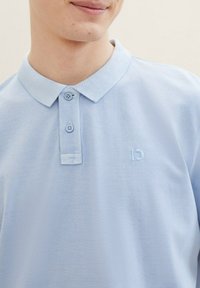 Ljust blå pikétröja med en texturerad krage, två knappar och en subtilt broderad logo på vänster bröst. Mjuk och andningsbar tyg.