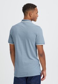 Bărbat stând cu spatele la cameră, purtând un tricou polo bleu cu mânecă scurtă și pantaloni închiși la culoare pe un fundal simplu.