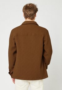 Mann in einer strukturierten braunen Jacke und weißen Hosen, der mit dem Rücken zur Kamera vor einem schlichten Hintergrund steht.