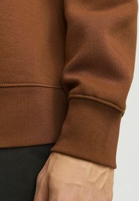 Tejido de sudadera marrón con puños acanalados, que presenta una textura suave y un diseño ajustado, mostrado junto a una mano parcialmente visible.