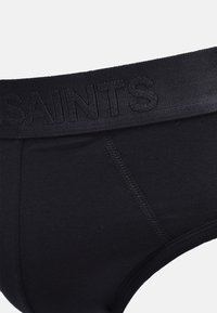 Sous-vêtements en coton noir avec une large ceinture élastique où est embossé le mot "SAINTS" en ton sombre. Texture lisse avec une coupe courbée.