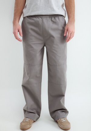 Man draagt losse grijze broek met elastische taille en beige veterschoenen, staand tegen een effen achtergrond.