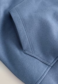 Gros plan sur un tissu de hoodie bleu clair, présentant une poche kangourou, une texture douce et des coutures visibles le long des bords et des coutures.