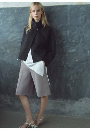 Veste noire en mélange de laine avec col haut, top en satin gris et shorts ajustés gris clair. Présente une texture douce et des lignes épurées.