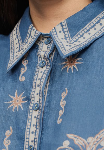 Μπλε πουκάμισο με κουμπιά και λευκά και μπεζ διακοσμητικά περιθώρια και μοτίβα ήλιου και στροβίλου πάνω στο ύφασμα, φορεμένο κάτω από το πηγούνι ενός ατόμου.