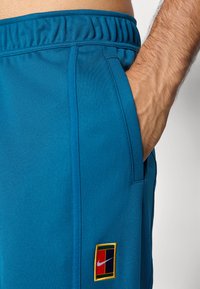 Artimiausias planas užsidariusios žmogaus rankos mėlynuose „Nike“ treninguose su elastingu juosmeniu ir daugiaspalviu logotipo u�žsegimu šalia kišenės.