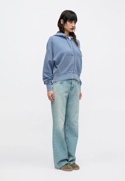 Μπλε βαμβακερό φούτερ με κουκούλα και τσέπη στο μπροστινό μέρος, συνδυασμένο με ανοιχτό μπλε τζιν παντελόνι φαρδιάς γραμμής και πράσινα σουέτ παπούτσια.