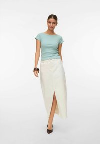 Vrouw in een lichtblauw aansluitend topje en een wit hoge taille rok met een split aan de voorkant, draagt donkere puntige hakken en bruine armbanden, staand tegen een witte achtergrond.