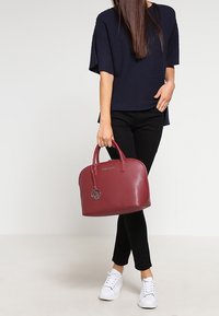 Bordó kožená kabelka s kulatými horními držadly, jemnou texturou a logovým přívěskem. Kombinováno s černým outfitem a bílými teniskami.