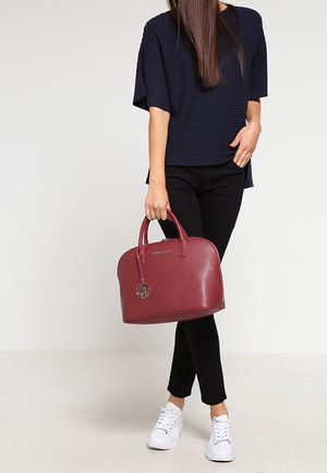 Sac à main en cuir bordeaux avec des poignées rondes, une texture légère et un bijou logo. Associé à une tenue noire et des baskets blanches.