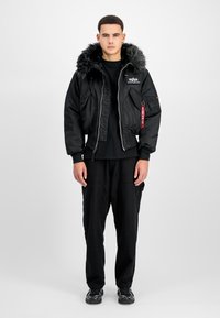 Schwarze Bomberjacke mit einem mit Fell gefütterten Kapuze, Reißverschluss vorne und Seitentaschen. Getragen über einem schwarzen Shirt und kombiniert mit schwarzen Hosen und Schuhen.