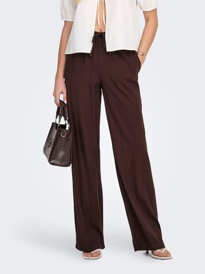 Femme portant un pantalon large marron foncé et un chemisier blanc ouvert devant, tenant un sac à main marron foncé, debout devant un fond uni.