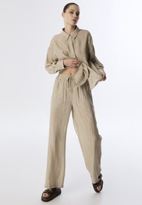 Camisa de lino beige con botones y cuello, combinada con pantalones sueltos a juego. Tejido texturizado con sutiles rayas verticales y una cinturilla elástica.