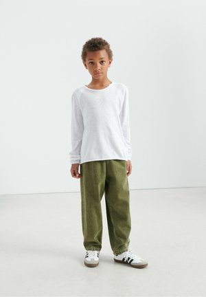 Balti ilgomis rankovėmis marškiniai su nesuformuota apačia, derinami su alyvuogių žalių laisvo silueto kelnėmis ir baltais sportbačiais su juodomis juostelėmis. Paprastas dizainas, kasdienis įvaizdis.