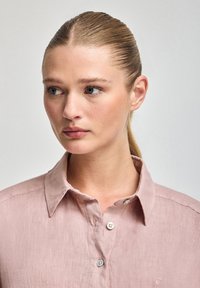 Camicia di lino rosa chiaro con colletto button-down, dotata di piccoli bottoni e una texture sottile. Il design include una tasca sul petto e un logo.